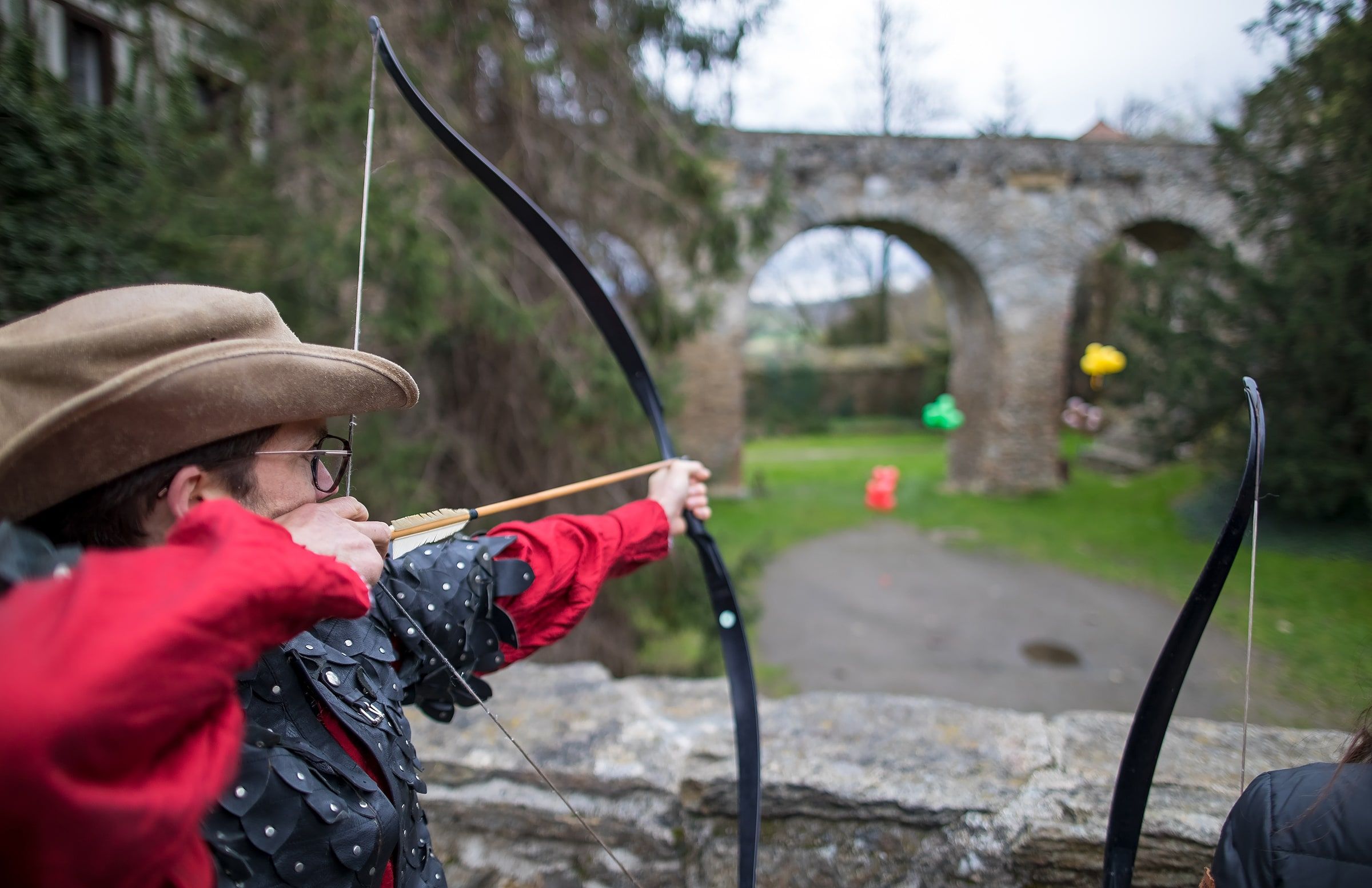 A player in a red coat and cowboy hat aims a bow at coloured balloons in front of a stone bridge