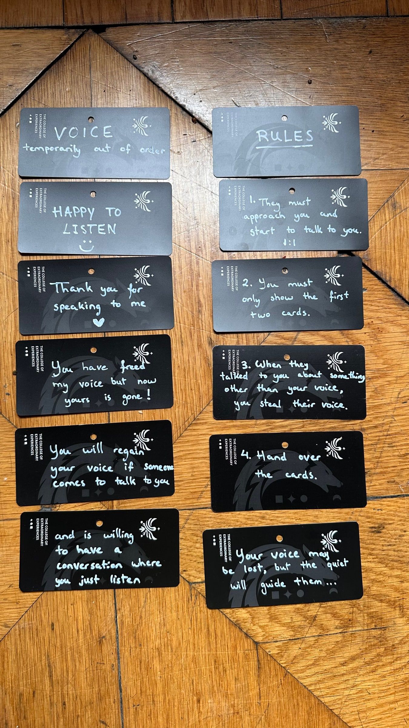 The twelve handwritten cards of the 2024 prototype laid out on a wooden floor, including VOICE temporarily out of order, HAPPY TO LISTEN and the four rules