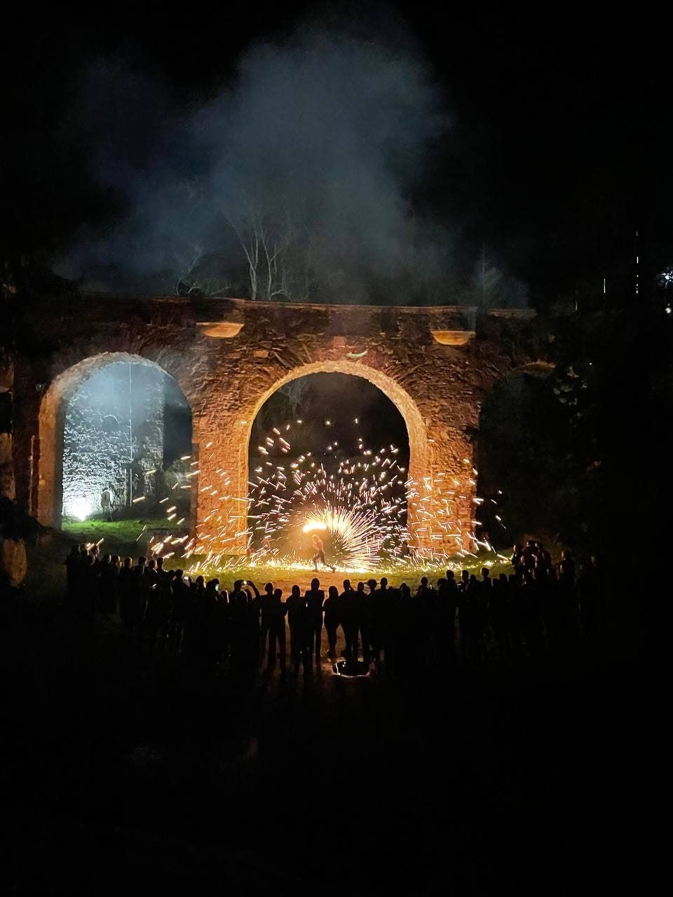 A fire performer spins a shower of sparks under a stone arch at night, watched by a crowd