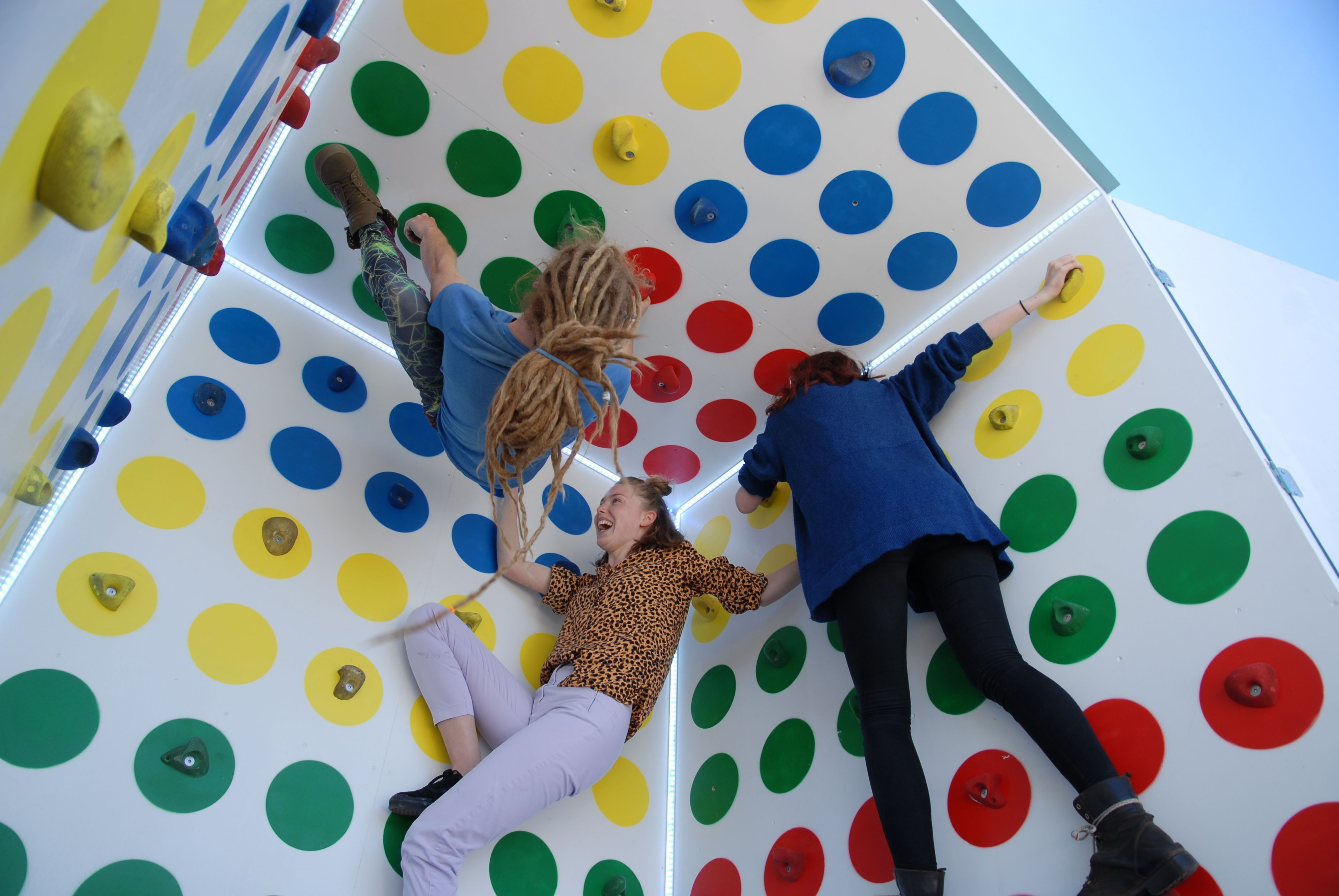 Three people climbing inside a white cube covered in red, yellow, green and blue dots with climbing holds