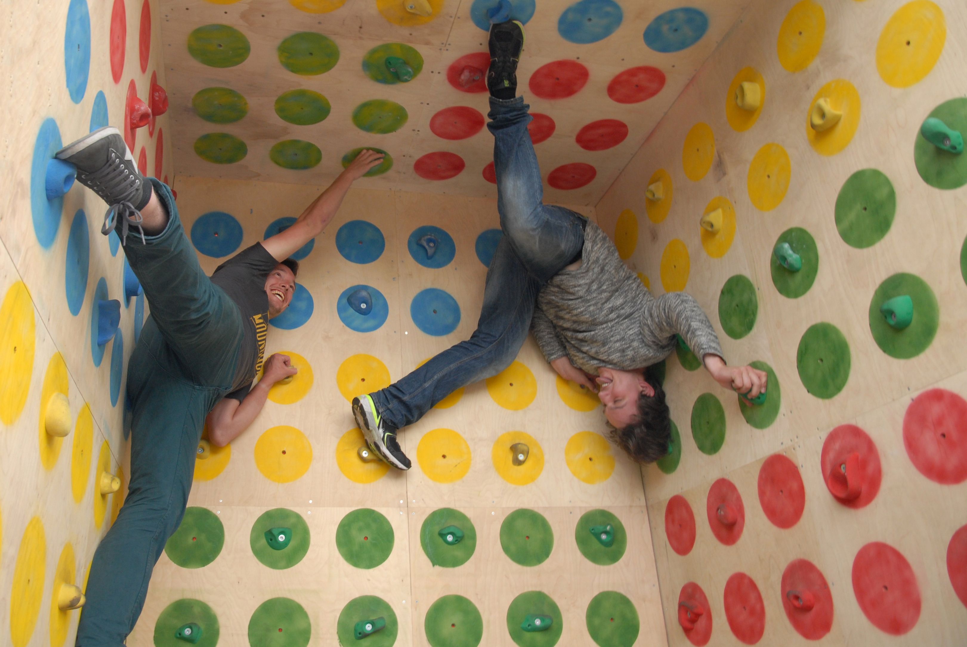 Two people hanging upside down in a wooden cube covered in coloured dots with climbing holds