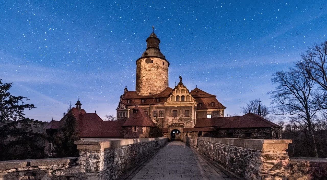 Castle Czocha at night under a starry sky, with its round stone tower and a bridge leading to the gate