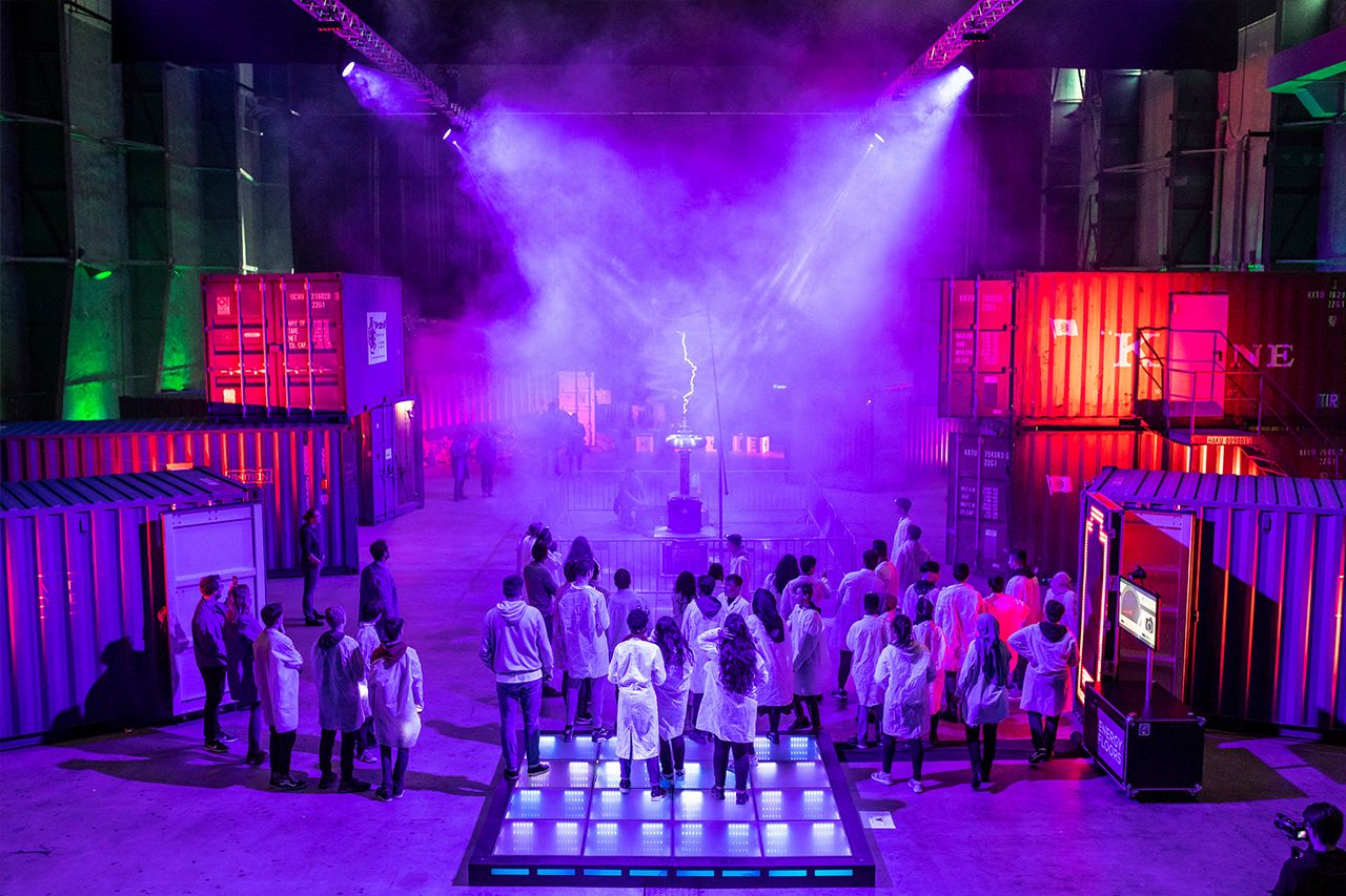 Students in white lab coats gather around a lit floor among shipping containers bathed in purple light