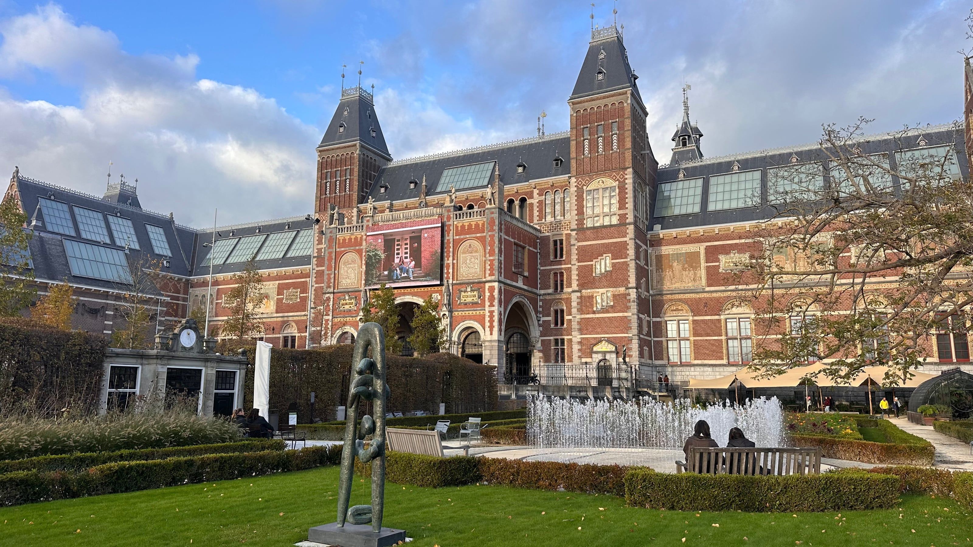 The Rijksmuseum in Amsterdam seen from its garden, with a fountain, a bronze sculpture and visitors on a bench in the autumn sun