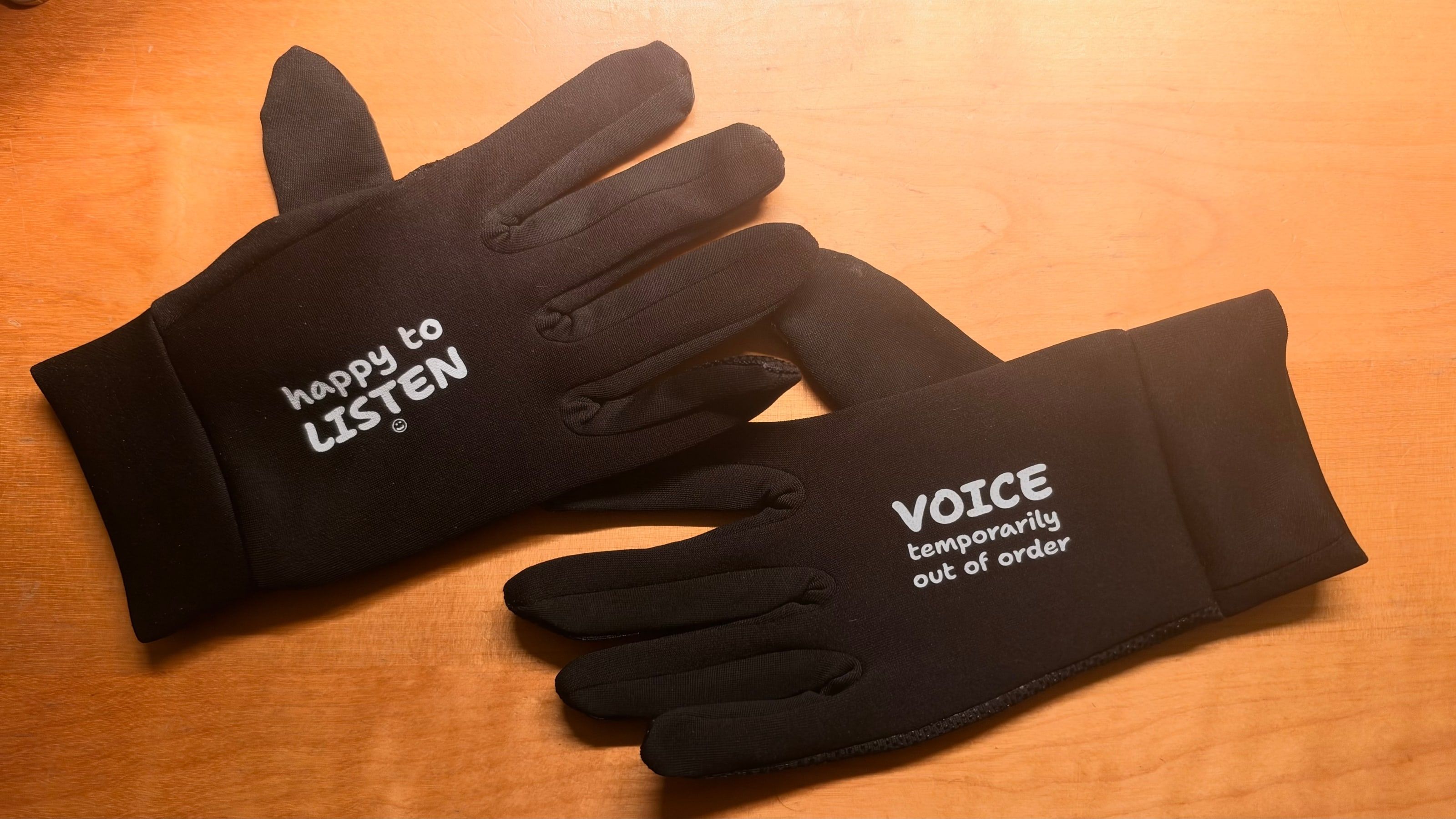 A pair of black gloves on a wooden table, printed with: happy to LISTEN and VOICE temporarily out of order